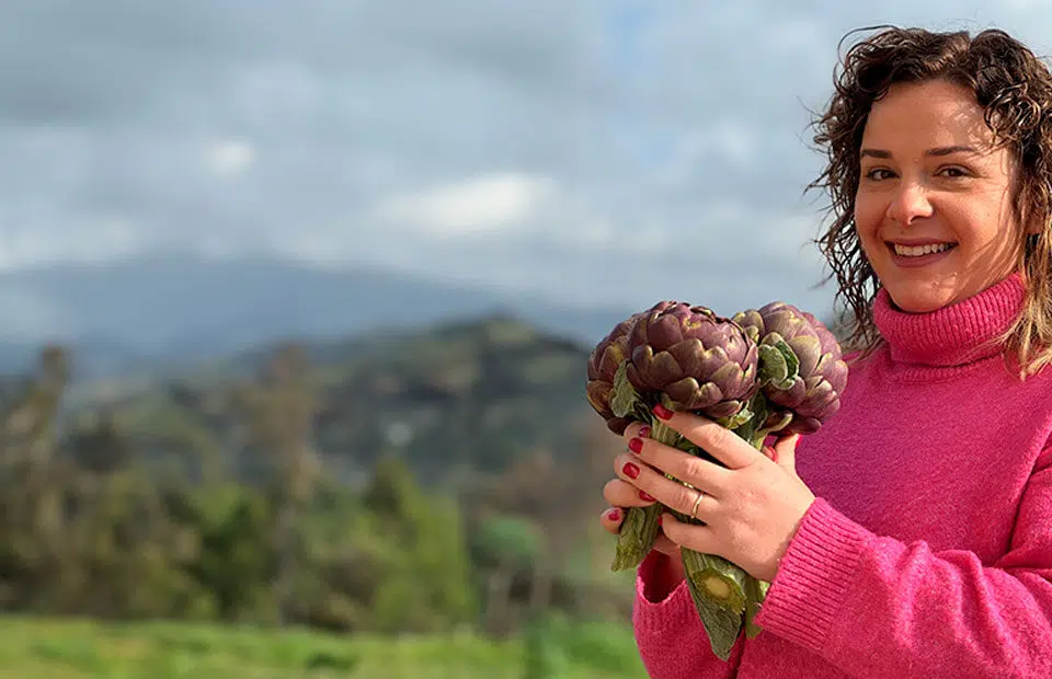 Panorama con Valentina a destra che indossa un maglione a collo alto fucsia mentre sorridente tiene in mano tre carciofi romaneschi