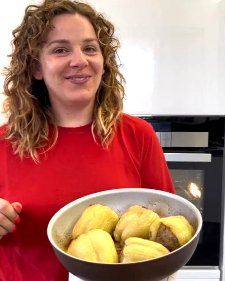 Ragazza bionda e riccia sorridente con maglia rossa che tiene in mano una teglia rotonda con 5 peperoni bianchi cotti e ripieni di carne